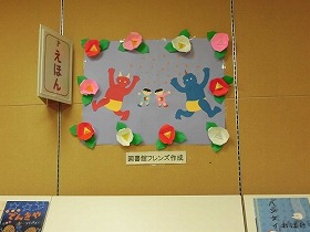 子供たちが豆まきをしている壁面装飾です。赤鬼と青鬼が一目散に逃げていきます。赤、白、ピンクのツバキの花がポスターの周りを囲んでいます。