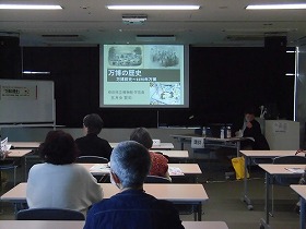 講演会が始まる直前の様子。参加者が席についている。正面のスクリーンにスライドを映しながら、講師が説明をするところ。